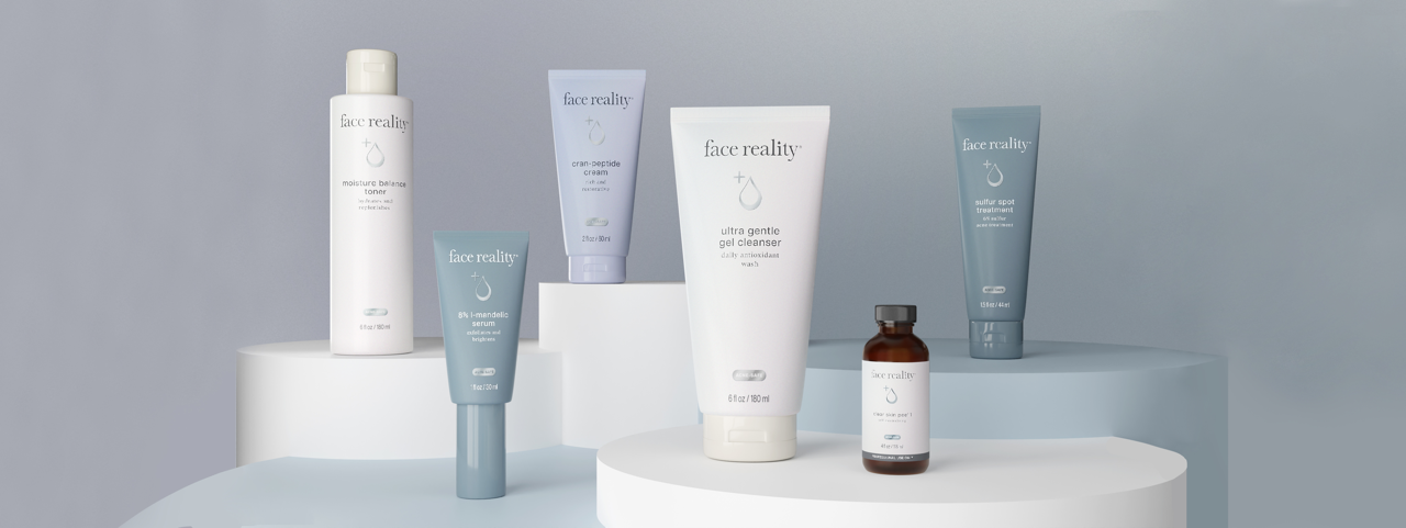Collection of Face Reality skincare products displayed on white curved surfaces, including various tubes, bottles, and jars in white, blue, and brown colors.