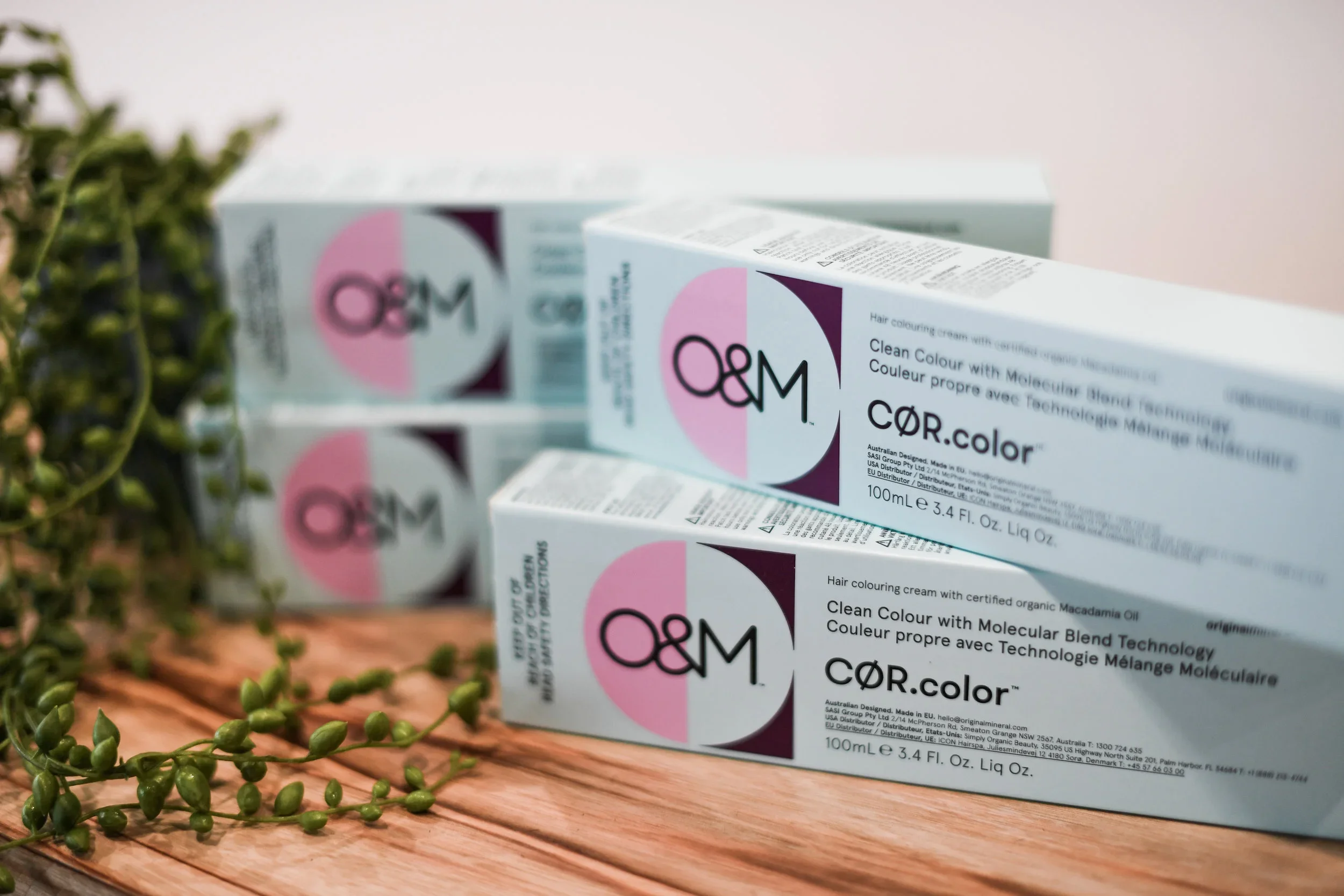 Three boxes of O&M CØR.color hair coloring cream on a wooden surface with green plant in the foreground.