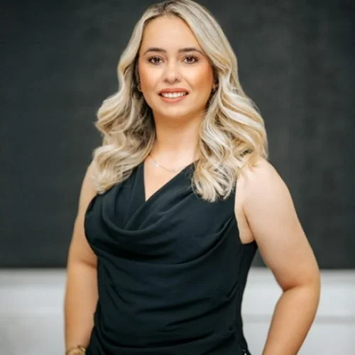A woman with long blonde hair wearing a sleeveless black dress, standing indoors against a dark background.