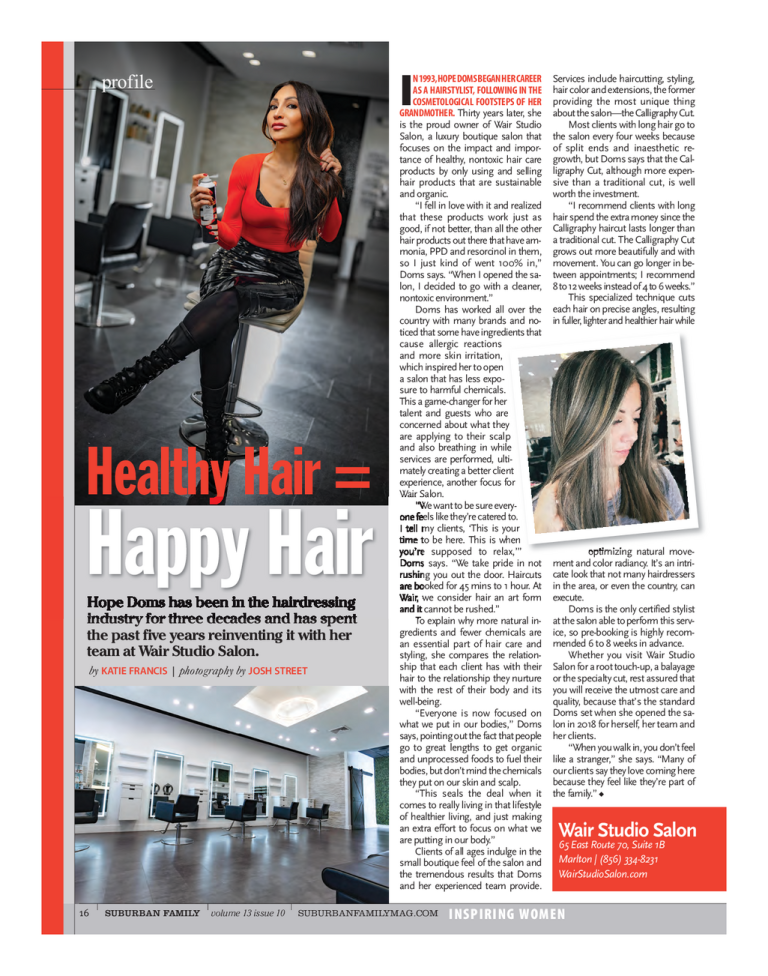 A woman in a red top and black leather skirt with lace-up boots sitting in a salon, spray bottle in hand. A second woman with long hair and glasses is seen in a mirror reflection, getting her hair styled.