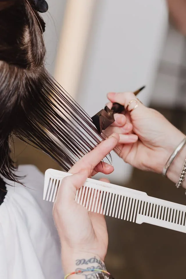 A hairstylist is cutting a woman's dark, straight hair with scissors, using a white comb to section the hair.