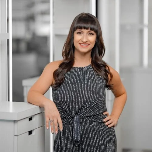 Woman with dark hair and bangs smiling, standing indoors with her hand on her hip, wearing a patterned sleeveless dress.