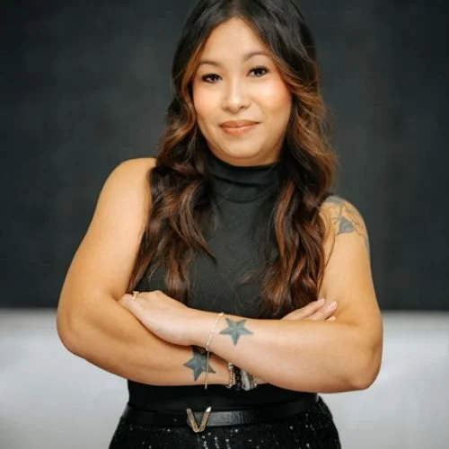 Woman with long wavy hair, wearing a sleeveless black top, crossing her arms, smiling, with tattoos on her arms, standing against a dark background.