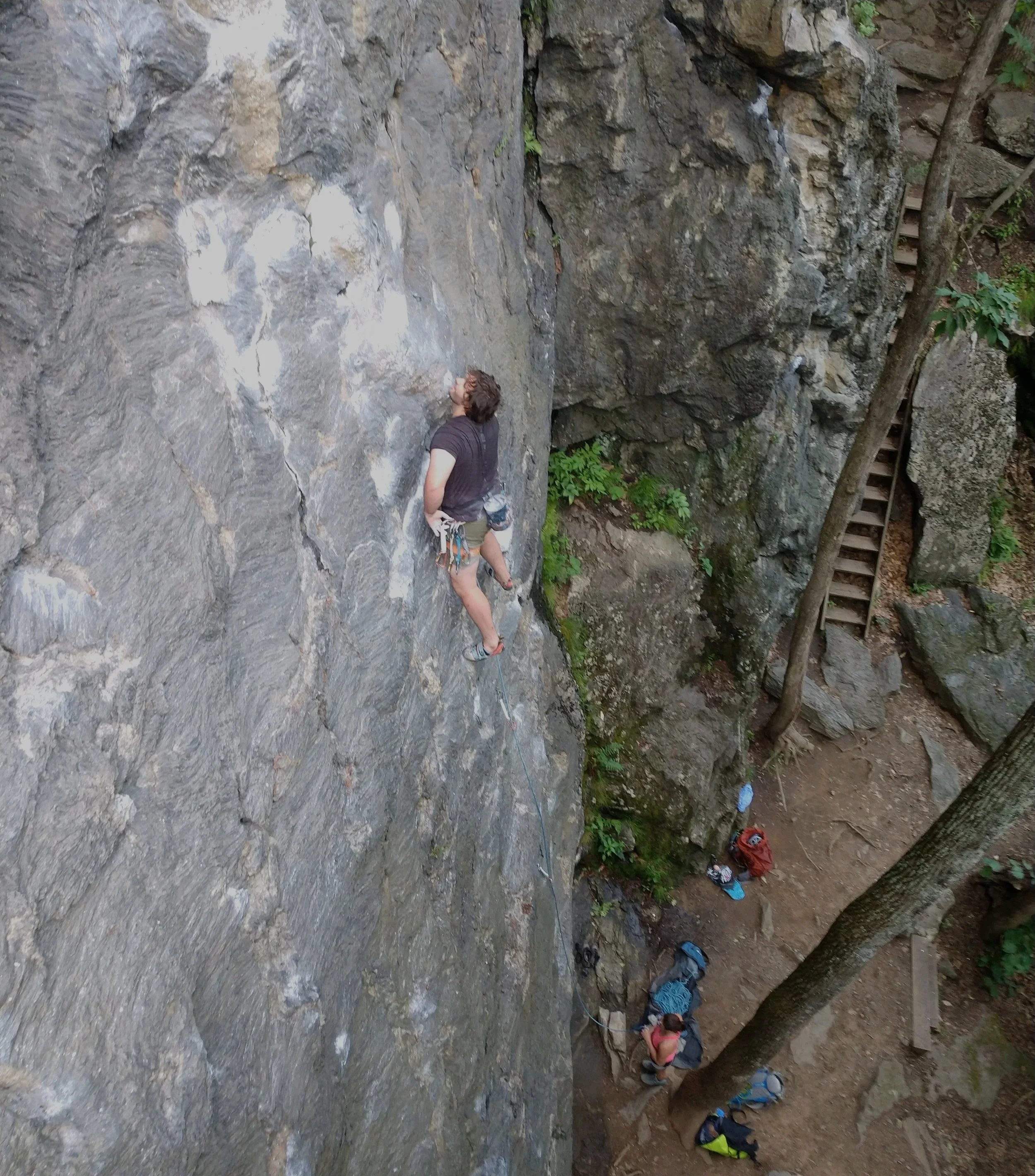 A person rock climbing on a steep outdoor cliff face with a harness and ropes, with another person below in a wooded area preparing to climb or belay.