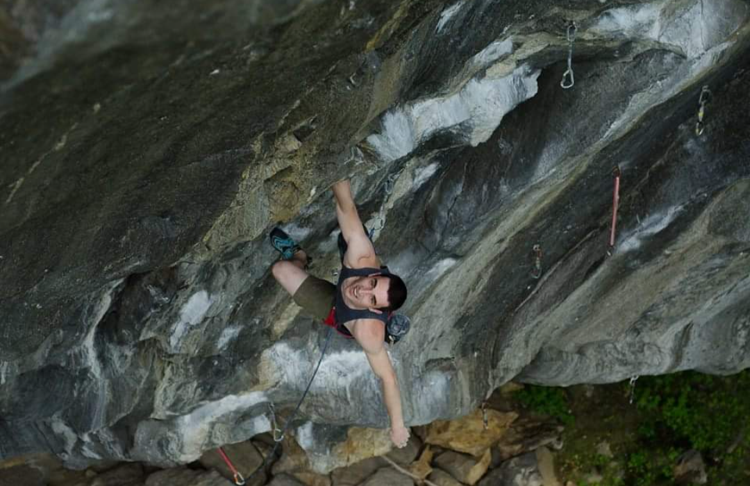 Person rock climbing on an overhanging cliff face, wearing a harness and climbing shoes, smiling and reaching for holds.