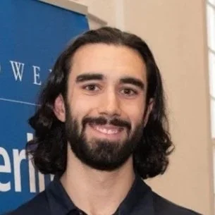 A man with long dark hair, a beard, and a mustache smiling, standing indoors in front of a blue backdrop.