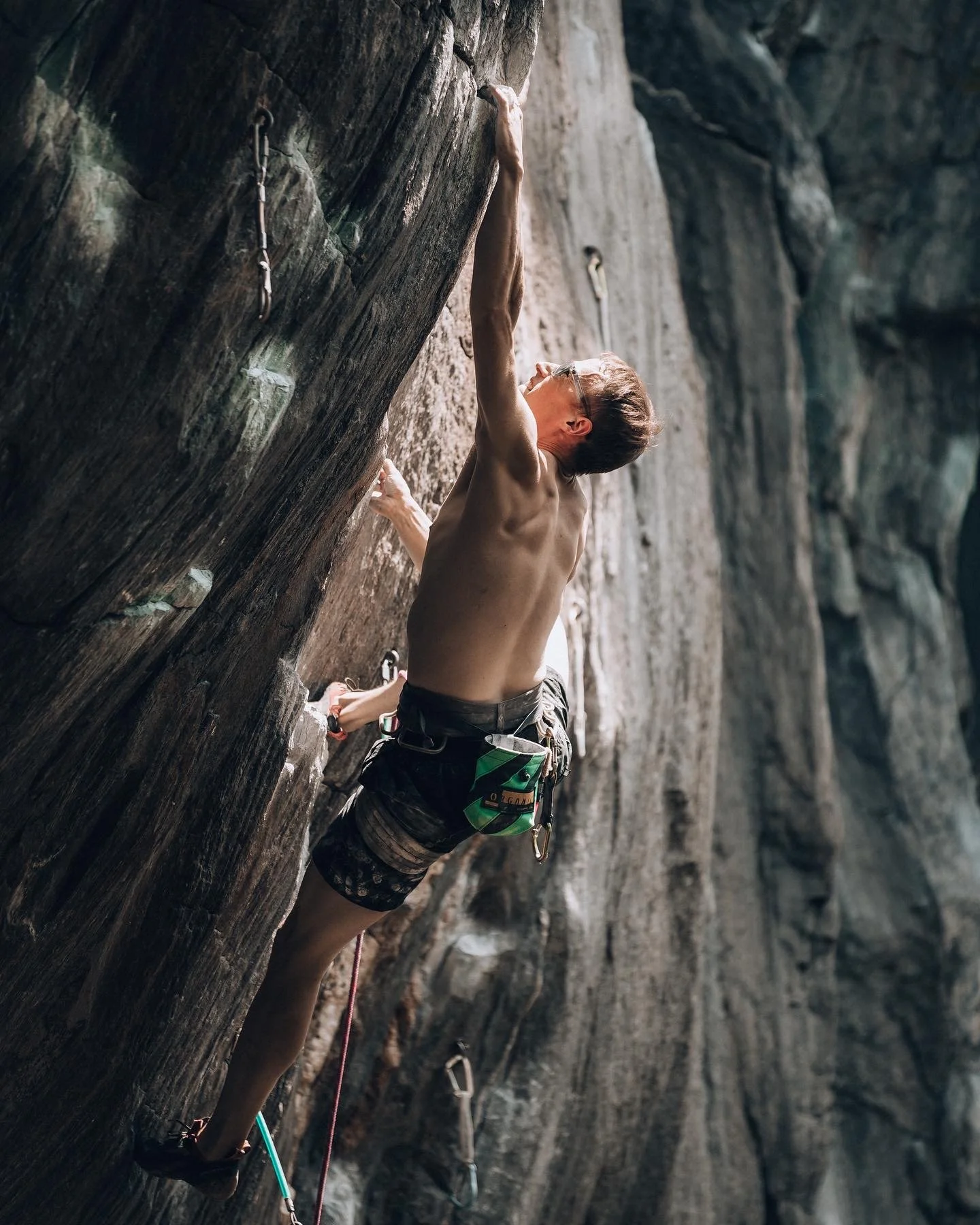 A shirtless male rock climber with glasses ascending a steep indoor climbing wall using harness and quickdraws.