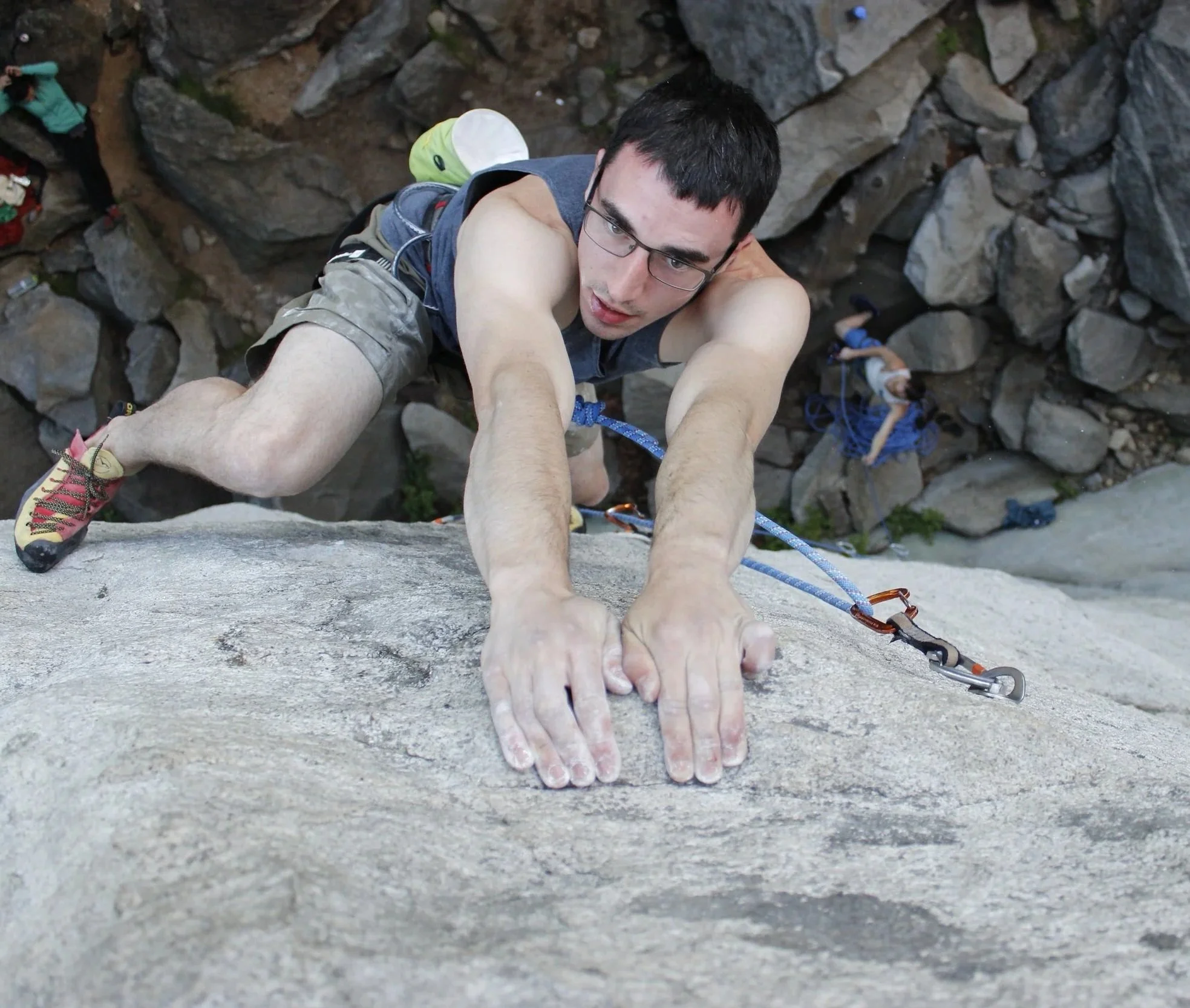 Climber ascending a rock face with quickdraw attached to his harness, with a belayer below.