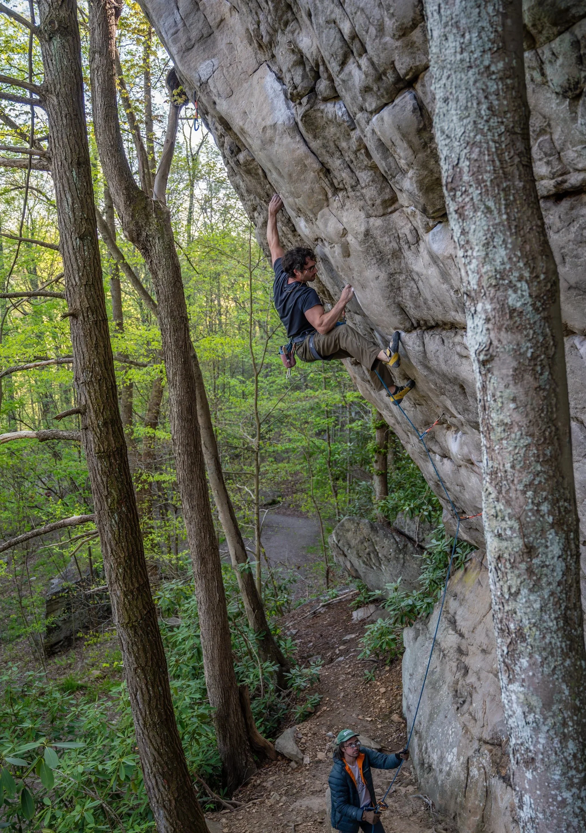 A man rock climbing on an outdoor cliff face, with a person below holding the safety rope in a wooded area.
