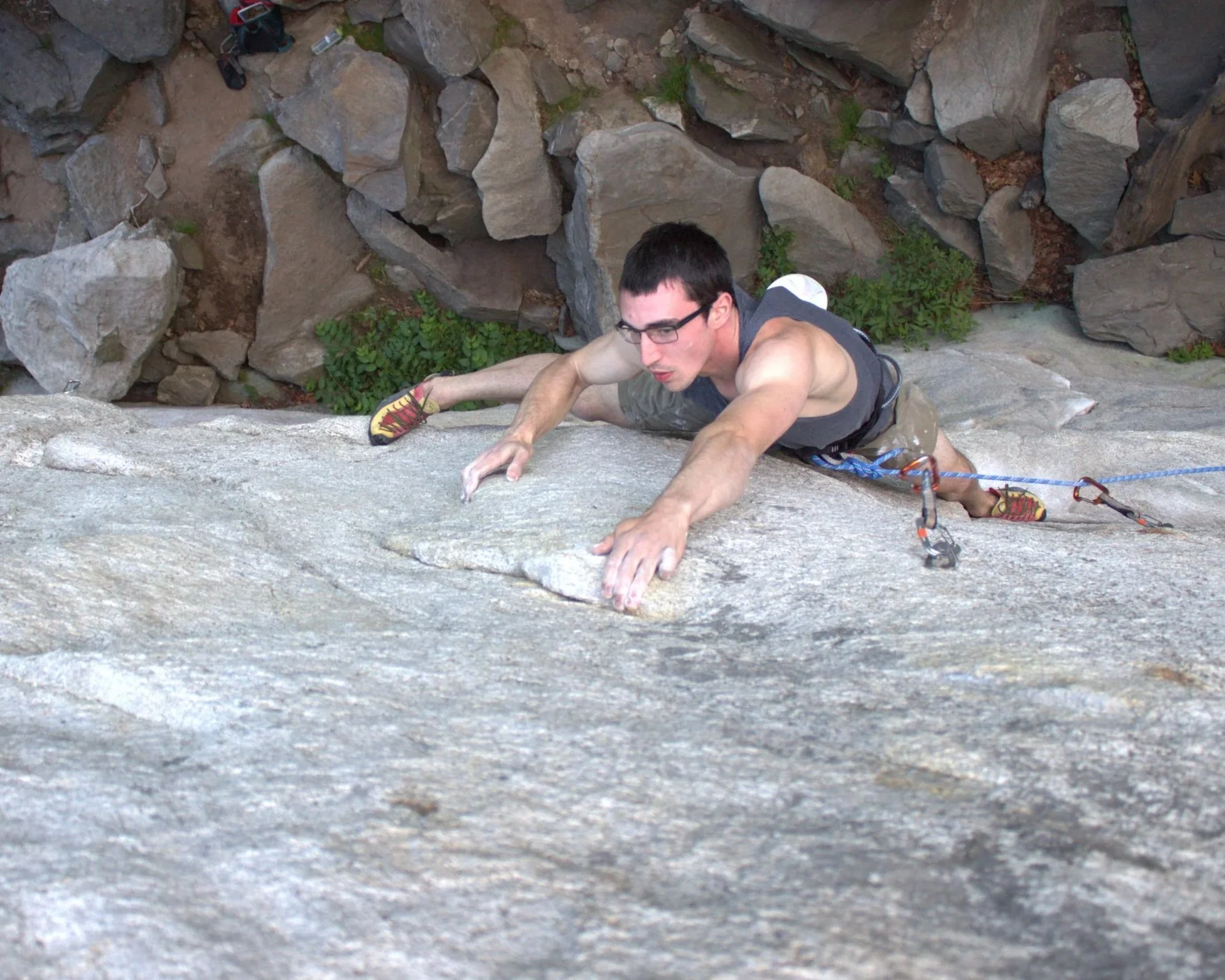 A man wearing glasses, a sleeveless shirt, and climbing shoes is rock climbing on a steep outdoor rock face, using his hands and feet for support, with a rocky and green background.