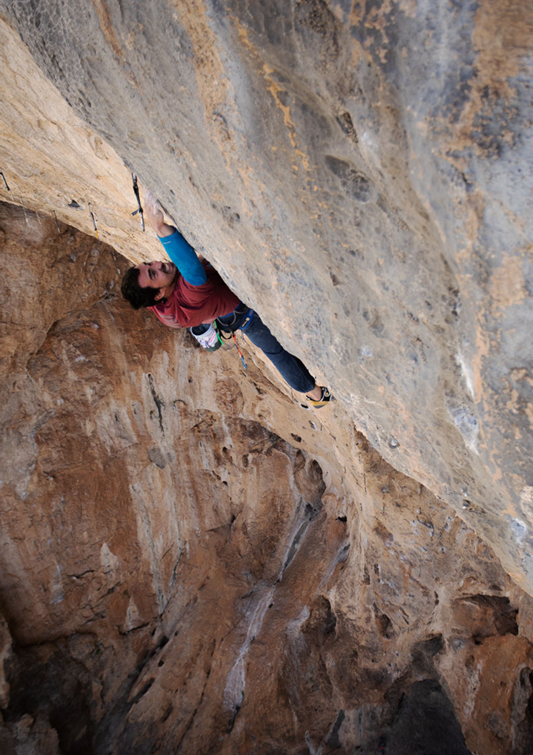 A man in climbing gear, wearing a red shirt and blue pants, is rock climbing on a steep outdoor rock face with a harness and carabiners, surrounded by rugged orange-brown rock formations.