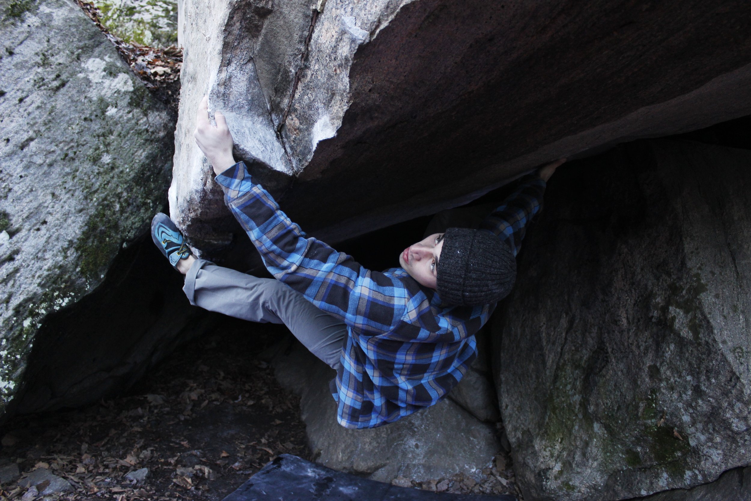 A person wearing a black beanie, a blue checkered jacket, and gray pants is bouldering on a large rock formation outdoors. The individual is reaching upward with one hand and has their legs bent, touching the rock with their feet.