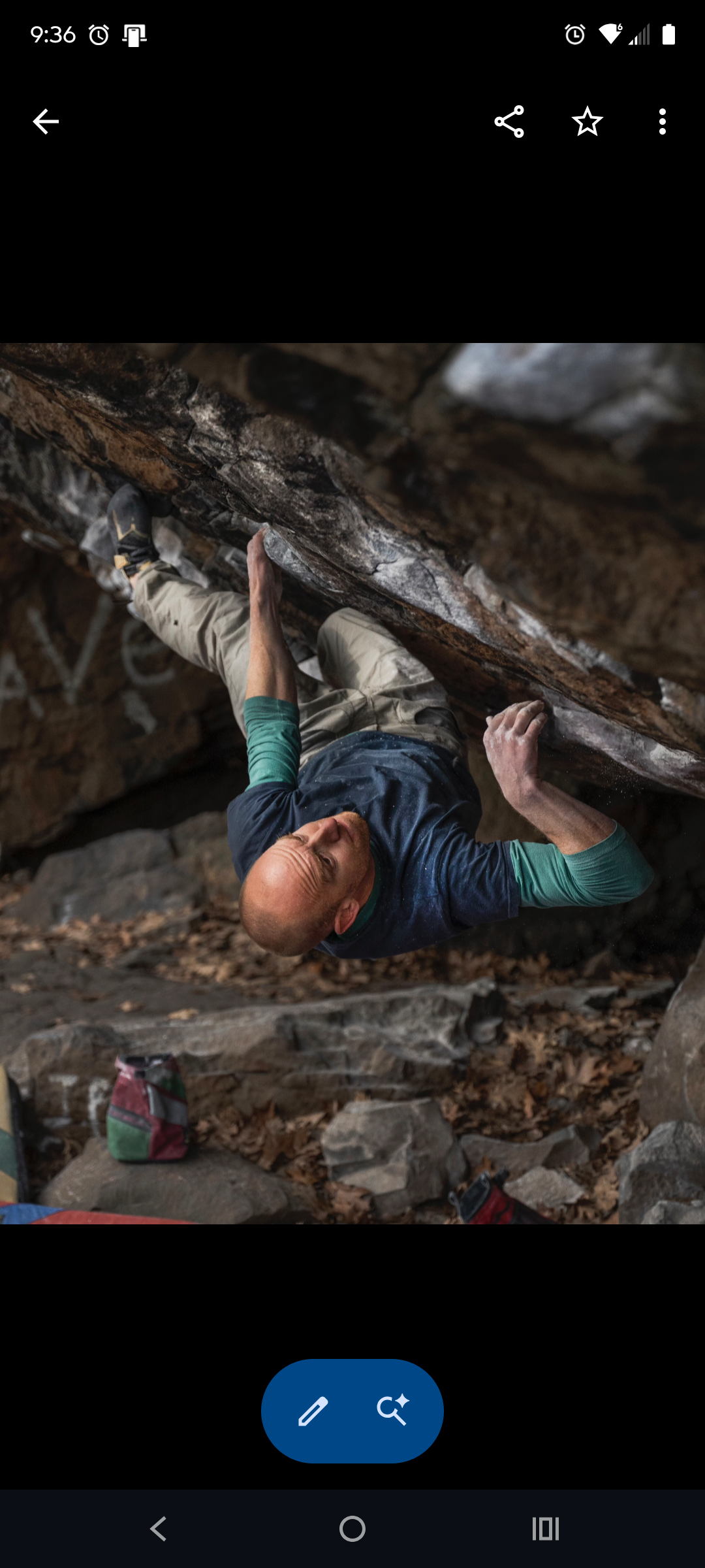 A man climbing a rock wall outdoors, wearing a blue shirt with green sleeves and beige pants, surrounded by rocks and fallen leaves.