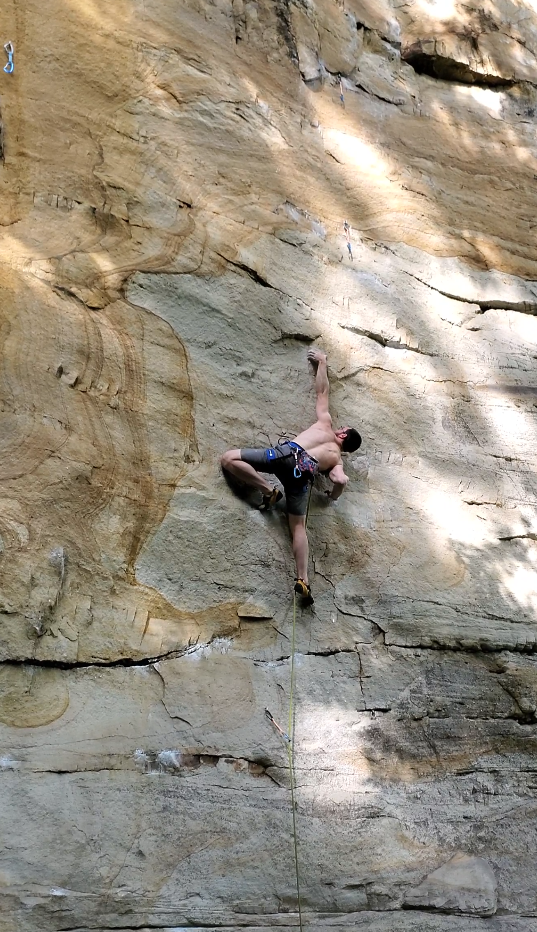 A shirtless man rock climbing on a vertical rock wall outdoors, wearing climbing harness and safety gear, with a climbing rope attached.