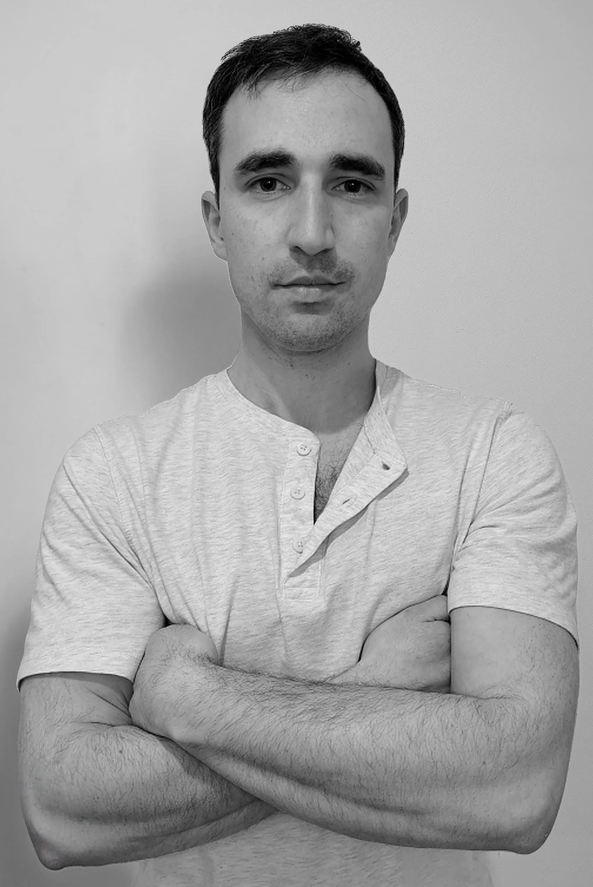 A black and white photo of a young man with short dark hair, wearing a light-colored, short-sleeved, button-up shirt, standing against a plain wall with arms crossed, looking directly at the camera.