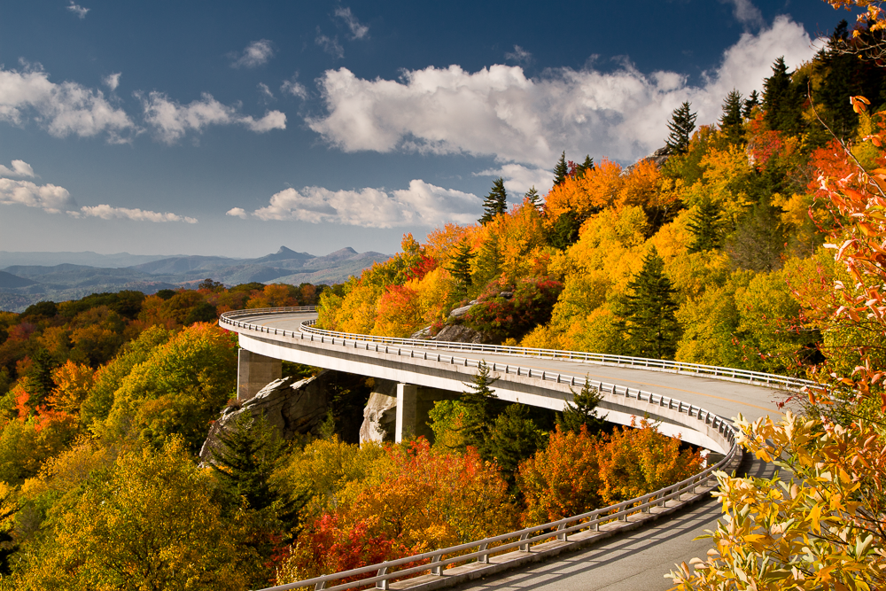 A winding mountain road surrounded by colorful fall foliage on a sunny day with blue sky and clouds.