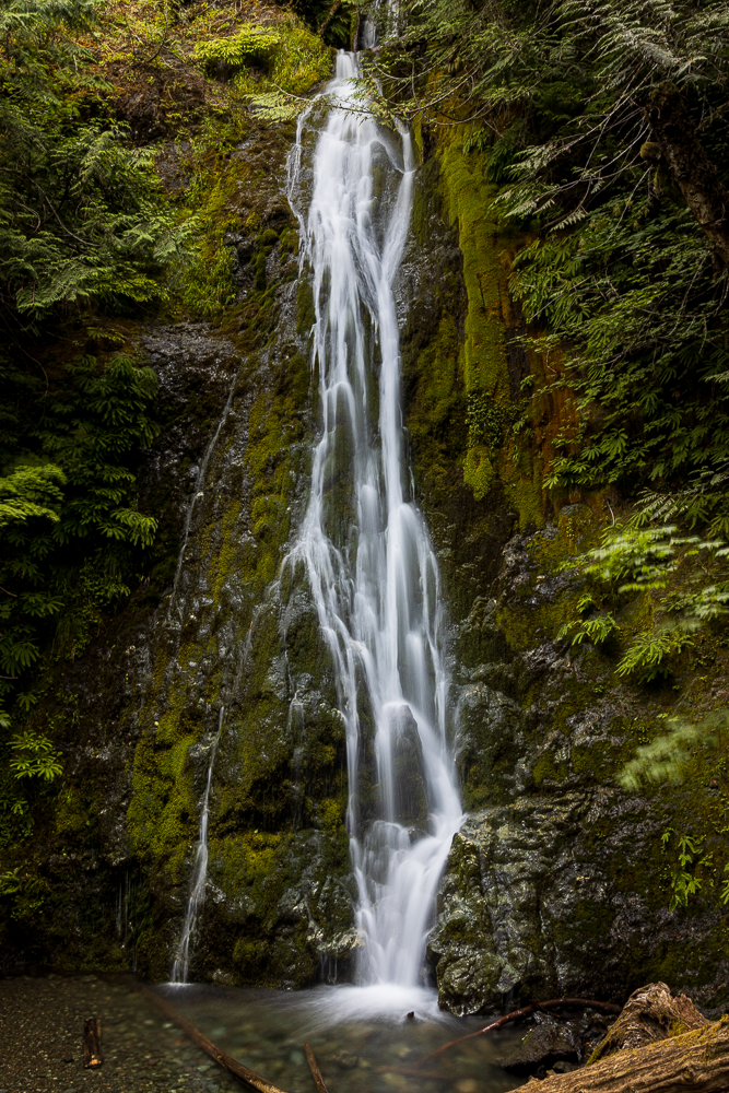 Tall waterfall cascading down moss-covered rocks surrounded by lush green forest.