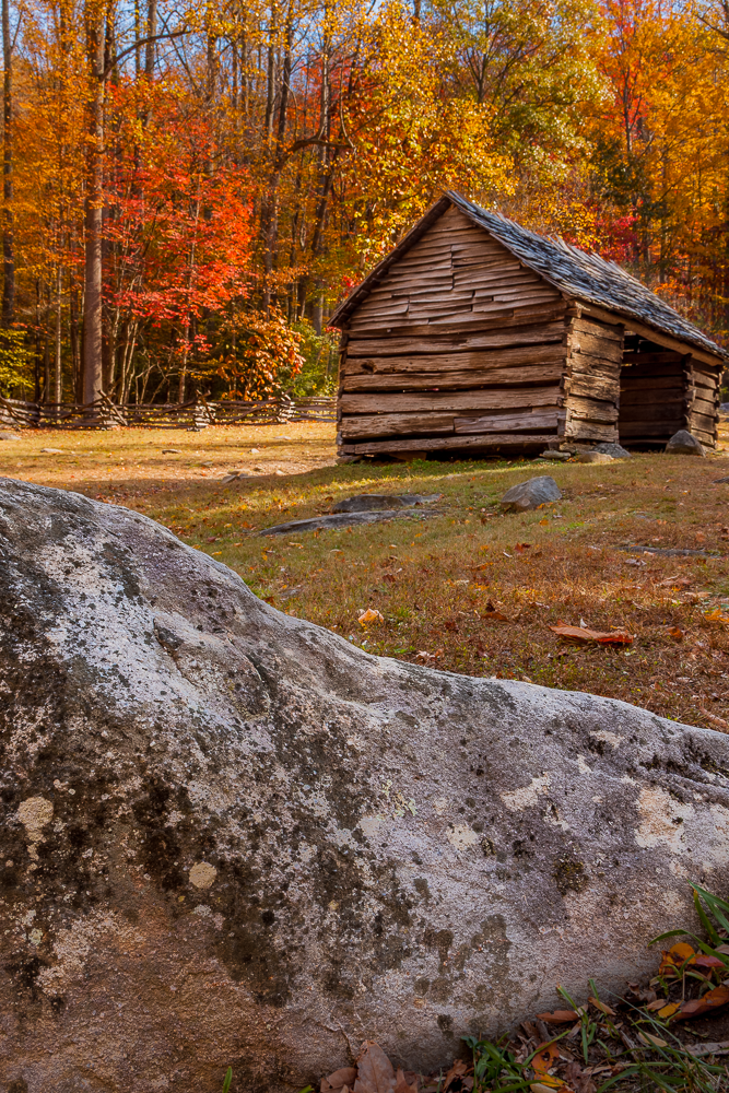 A rustic wooden cabin in an autumn forest with colorful fall foliage, a stone in the foreground, and a wooden fence in the background.