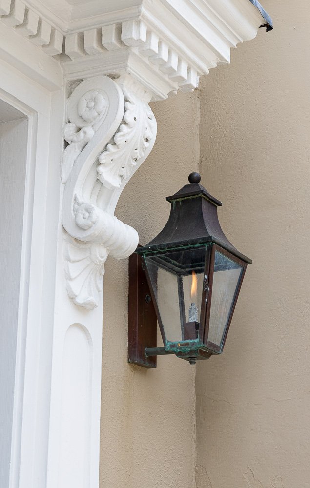 Close-up of a vintage black outdoor wall lantern with glass panels mounted on beige wall next to ornate white architectural molding and cornice.