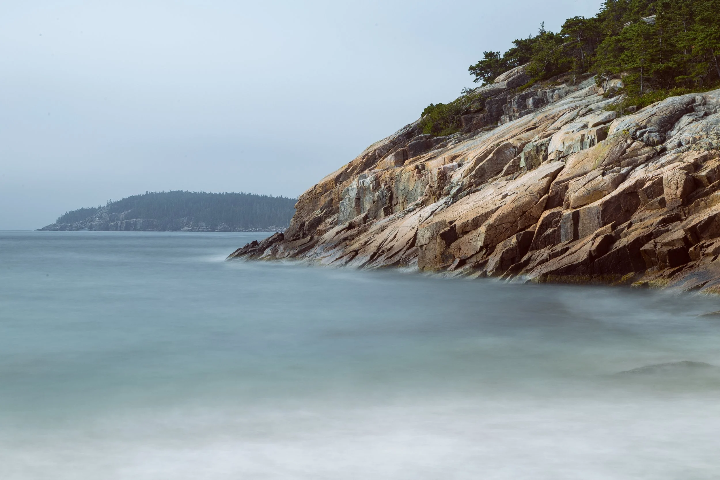 Coastal scene with rocky cliffs on the right and calm ocean waters in the foreground, with a forested island in the distance under a cloudy sky.