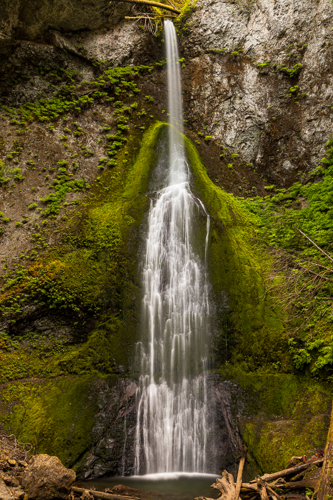 Tall waterfall flowing down a moss-covered rocky cliff in a lush forest setting.