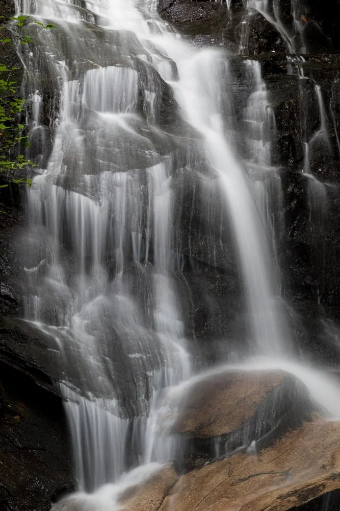 Long exposure photo of a multi-tiered waterfall cascading down dark rocks surrounded by green foliage.