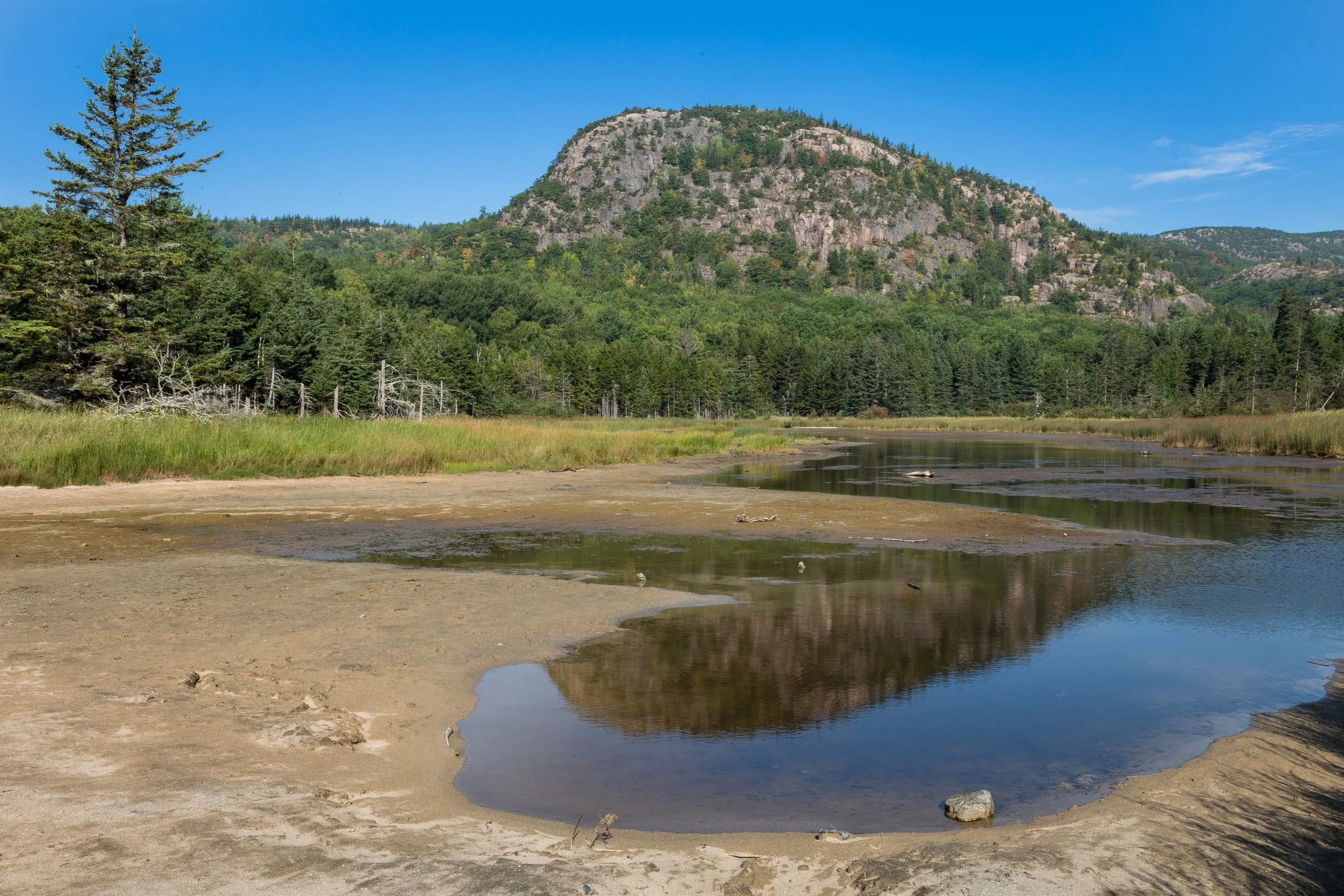 A scenic landscape showing a mountain with rocky cliffs, surrounded by green trees, and a river or pond in the foreground under a clear blue sky.