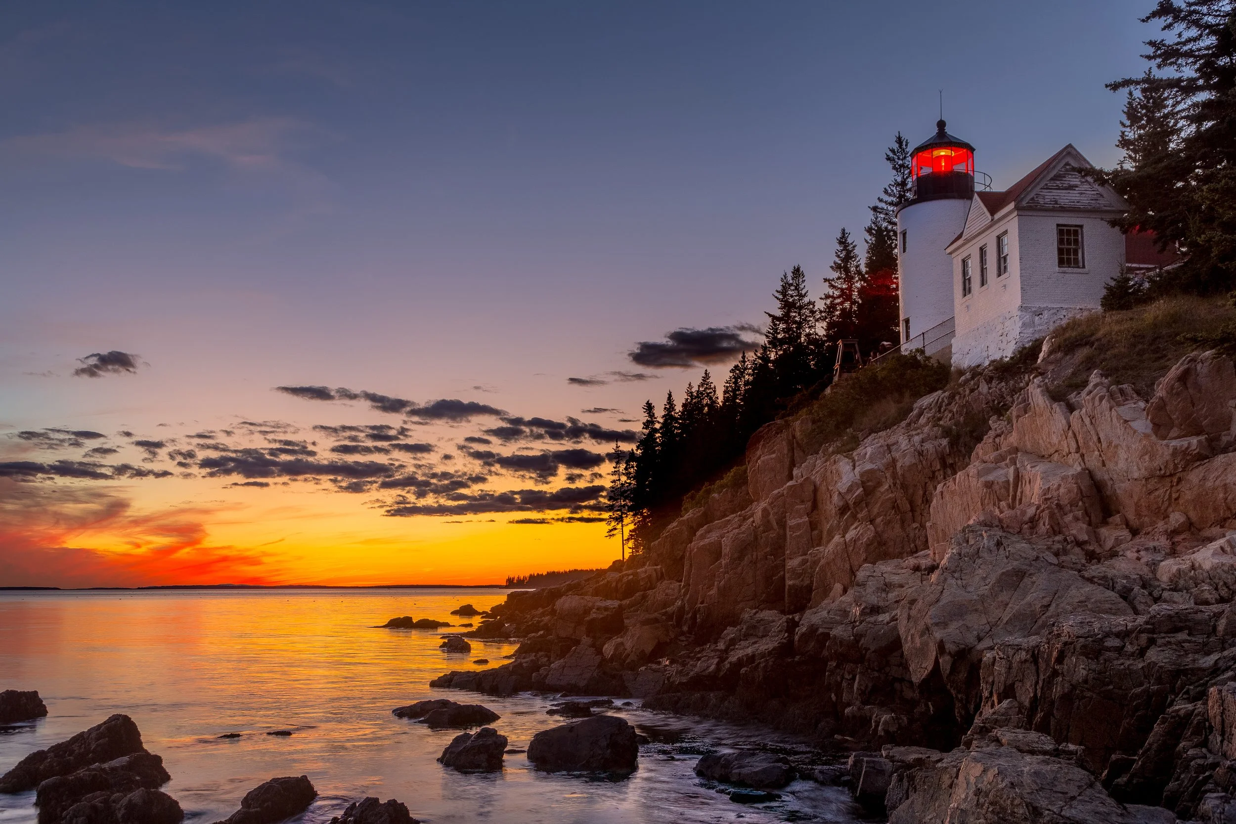 A lighthouse on a rocky coast at sunset with a partly cloudy sky.