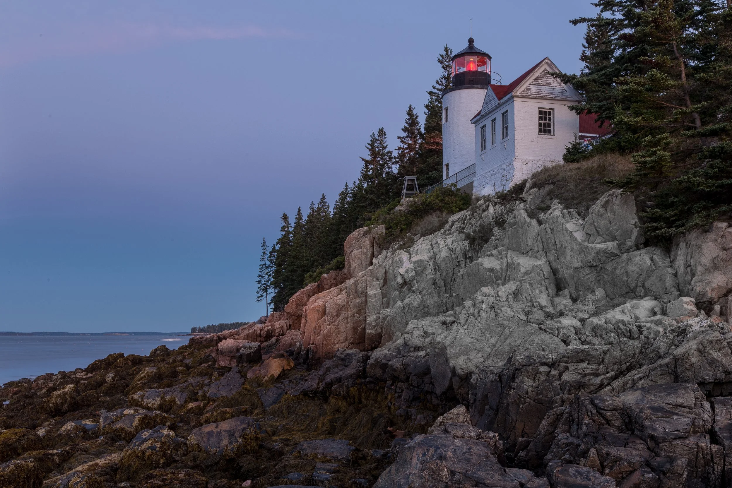 A lighthouse perched on a rocky cliff by the ocean with trees around it during twilight.