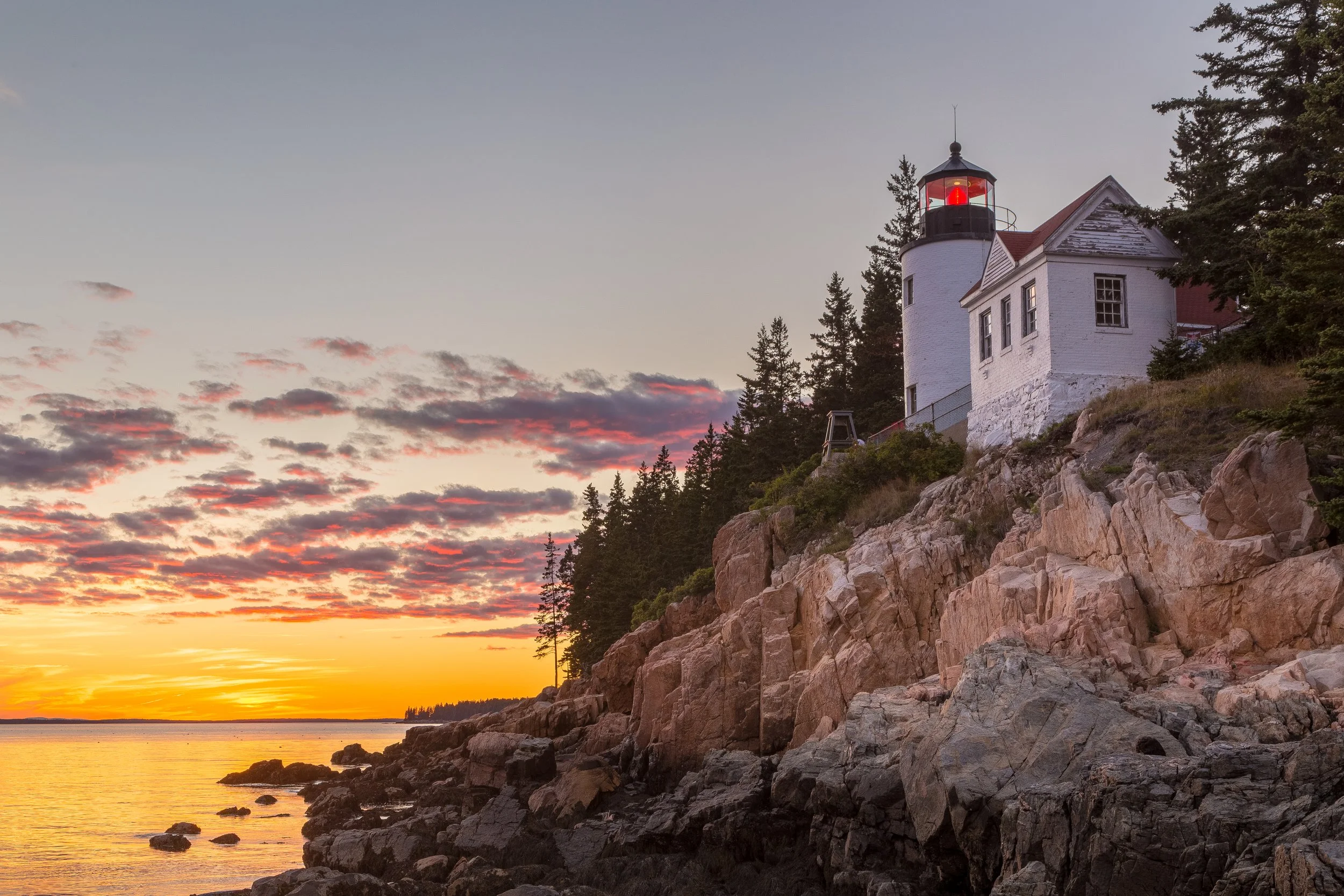 A lighthouse on a rocky coast at sunset with a partly cloudy sky and trees in the background.