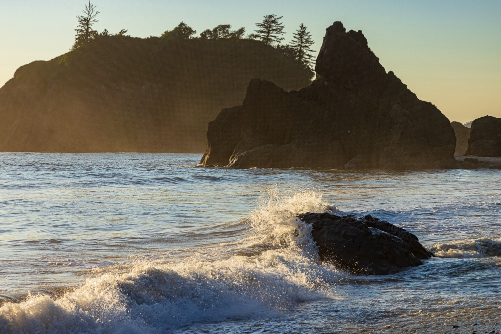 Sunset over a rocky shoreline with waves crashing and a large rock formation in the ocean, with trees on a hill in the background.