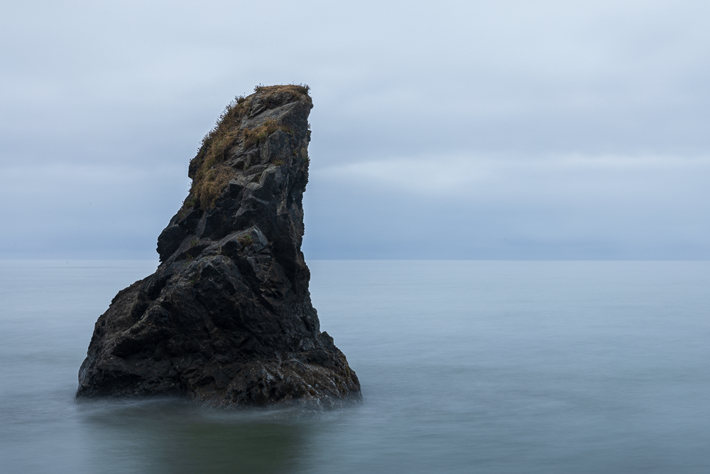 A large, dark rock formation in the ocean with a grassy top, under a cloudy sky.