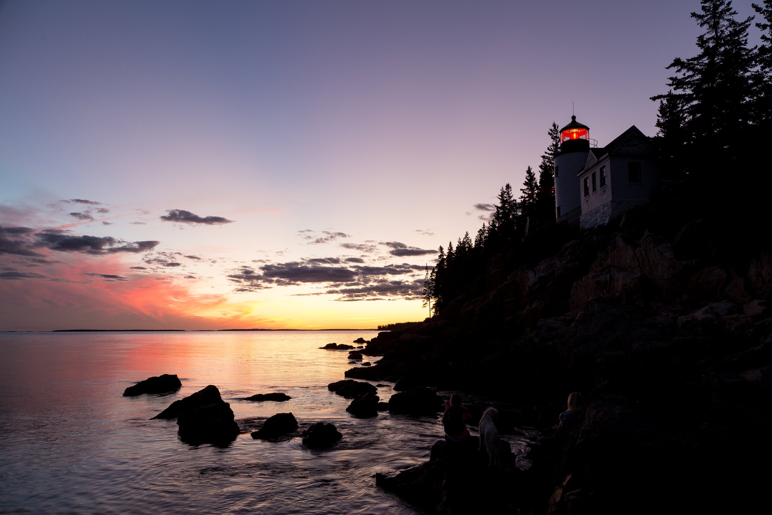Sunset over a body of water with rocks in the foreground, silhouette of people sitting by the shore, and a lighthouse on a hill surrounded by trees on the right.