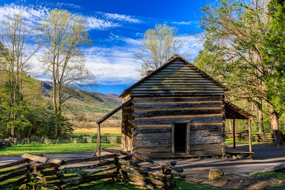 An old wooden barn in a rural setting, surrounded by trees and a split-rail fence, with mountains and a partly cloudy sky in the background.