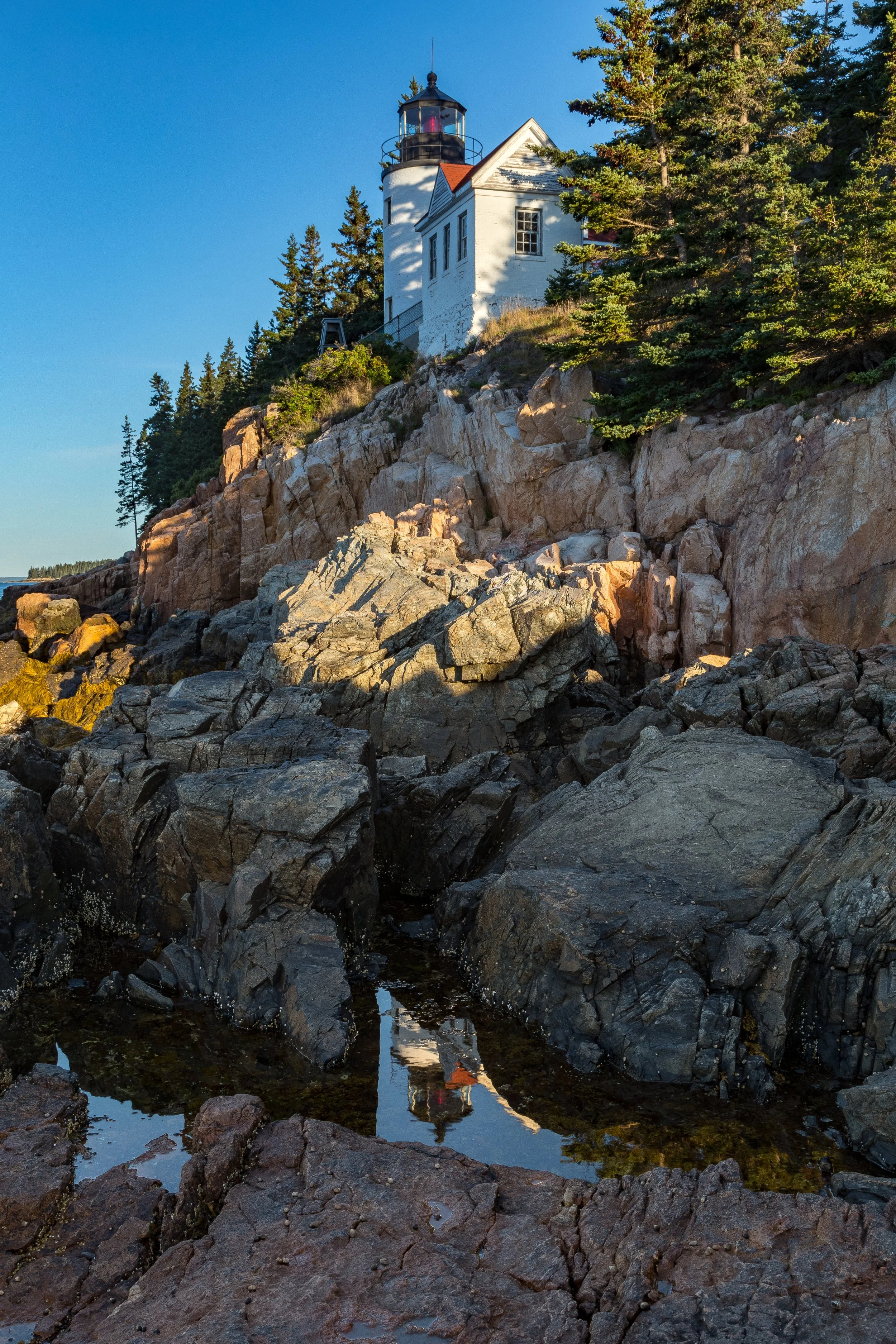 A lighthouse on a rocky shoreline with trees, reflected in a small pool of water in the foreground.