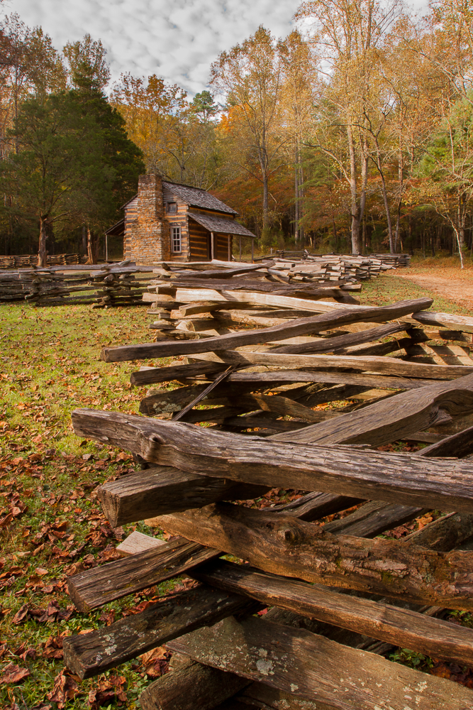 Old wooden fence leading to a small log cabin in a fall forest with colorful trees and a cloudy sky.
