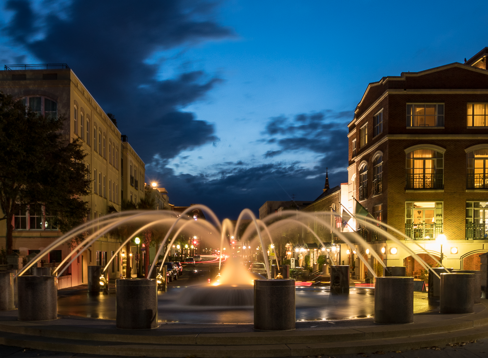 Night view of a city street with illuminated fountain in the center, surrounded by historic buildings with warm lighting, under a dark blue sky with clouds.
