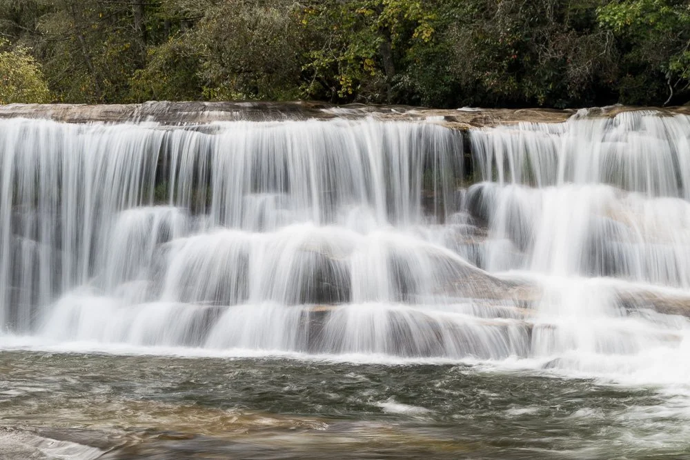 A waterfall flowing over rocks into a river, surrounded by trees.