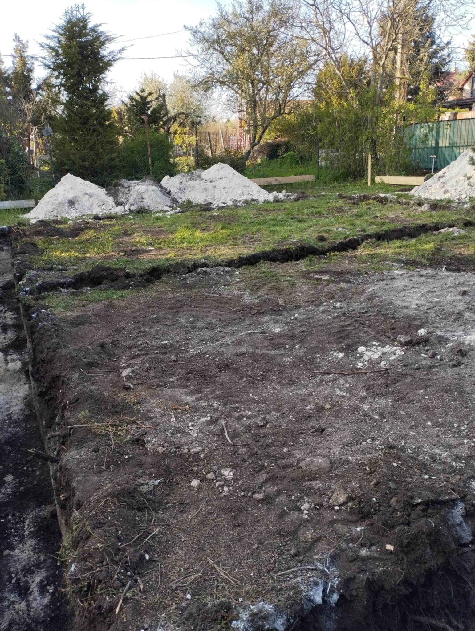 A backyard with patches of grass and dirt, multiple piles of white gravel or sand, and a background of trees and fences.