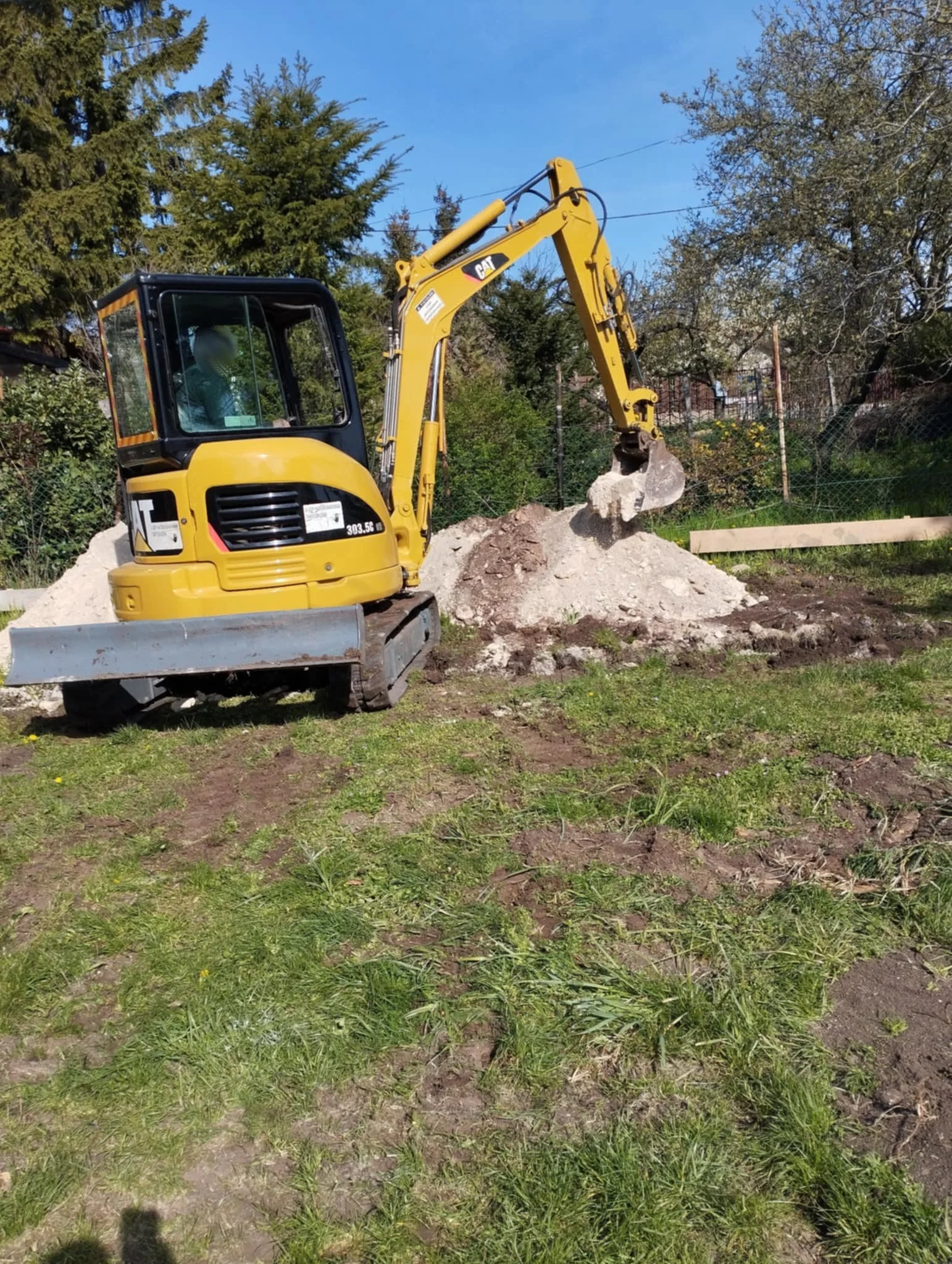 A yellow and black mini excavator scooping dirt at a construction site with green grass and trees in the background.