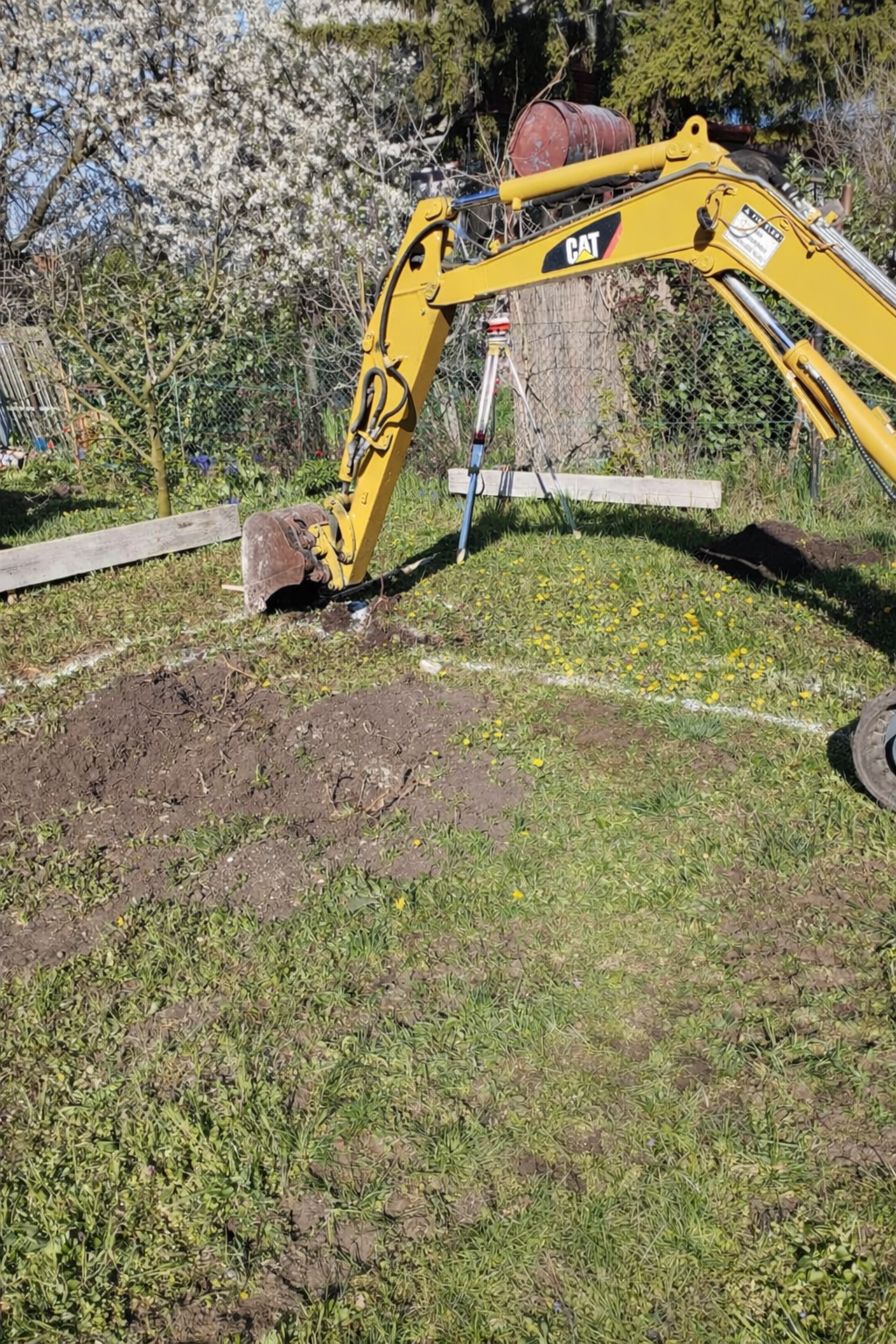 A yellow Caterpillar mini excavator with extended arm digging a hole in a grassy yard, with a wheelbarrow on top of the arm and a tripod with a leveling tool nearby. There are trees and a fence in the background.