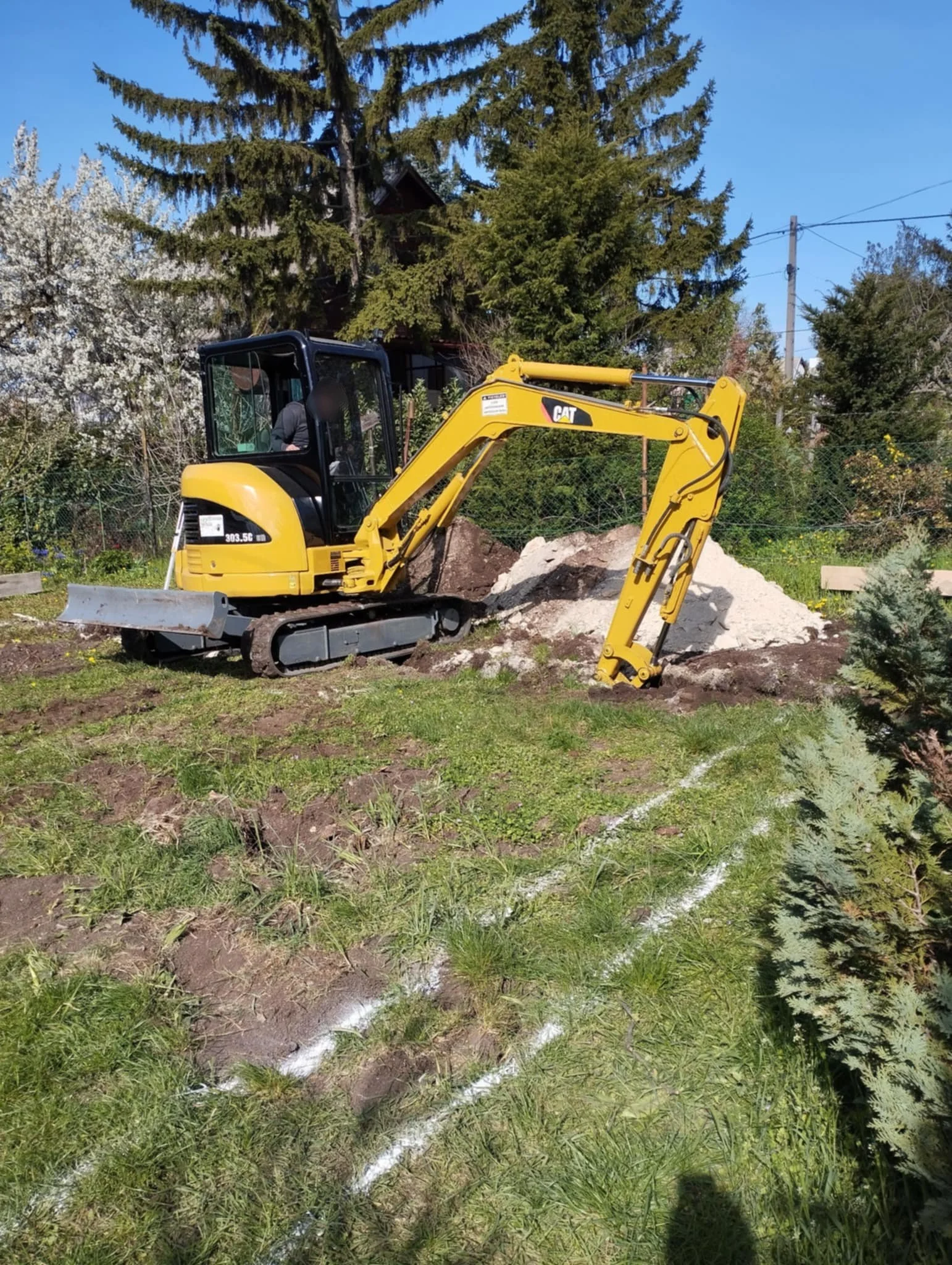 A small yellow excavator working on a garden or yard, with a pile of dirt and rocks nearby, under a bright blue sky with trees and a house in the background.