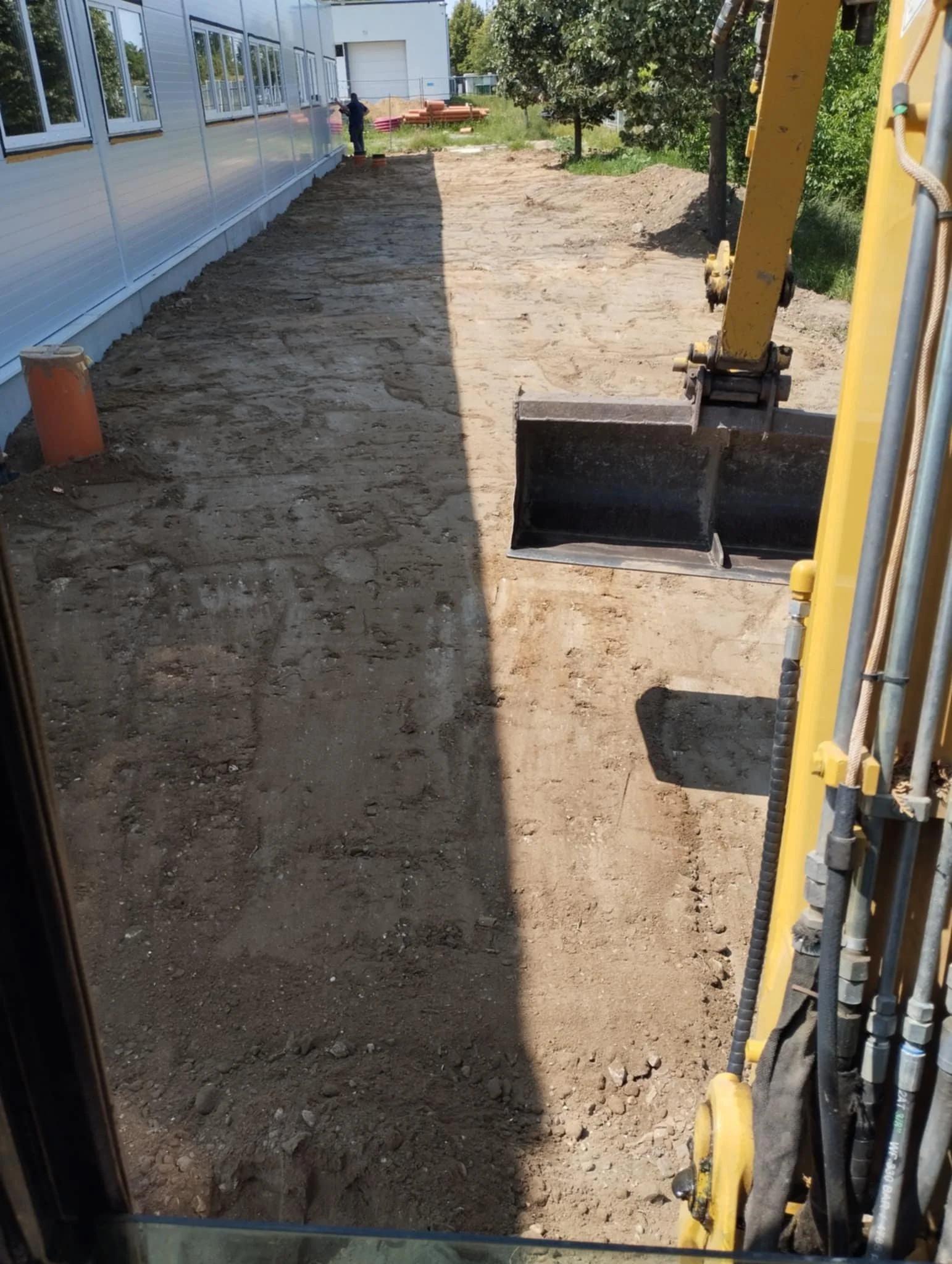 Construction site with cleared dirt area next to a building, a small excavator on the right, and people working in the background near trees and building materials.