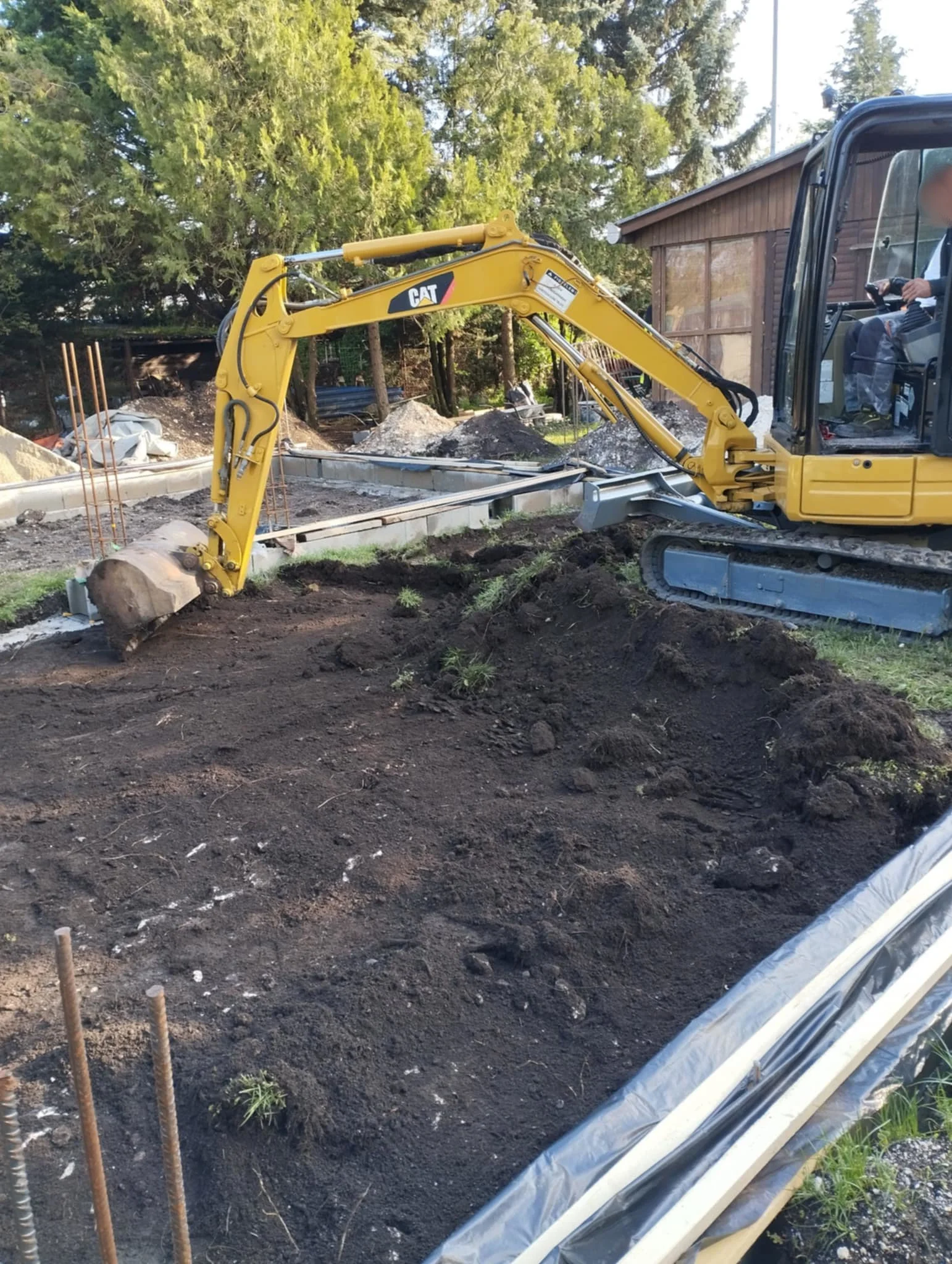 A yellow CAT excavator is digging soil at a construction site with trees and a wooden shed in the background.