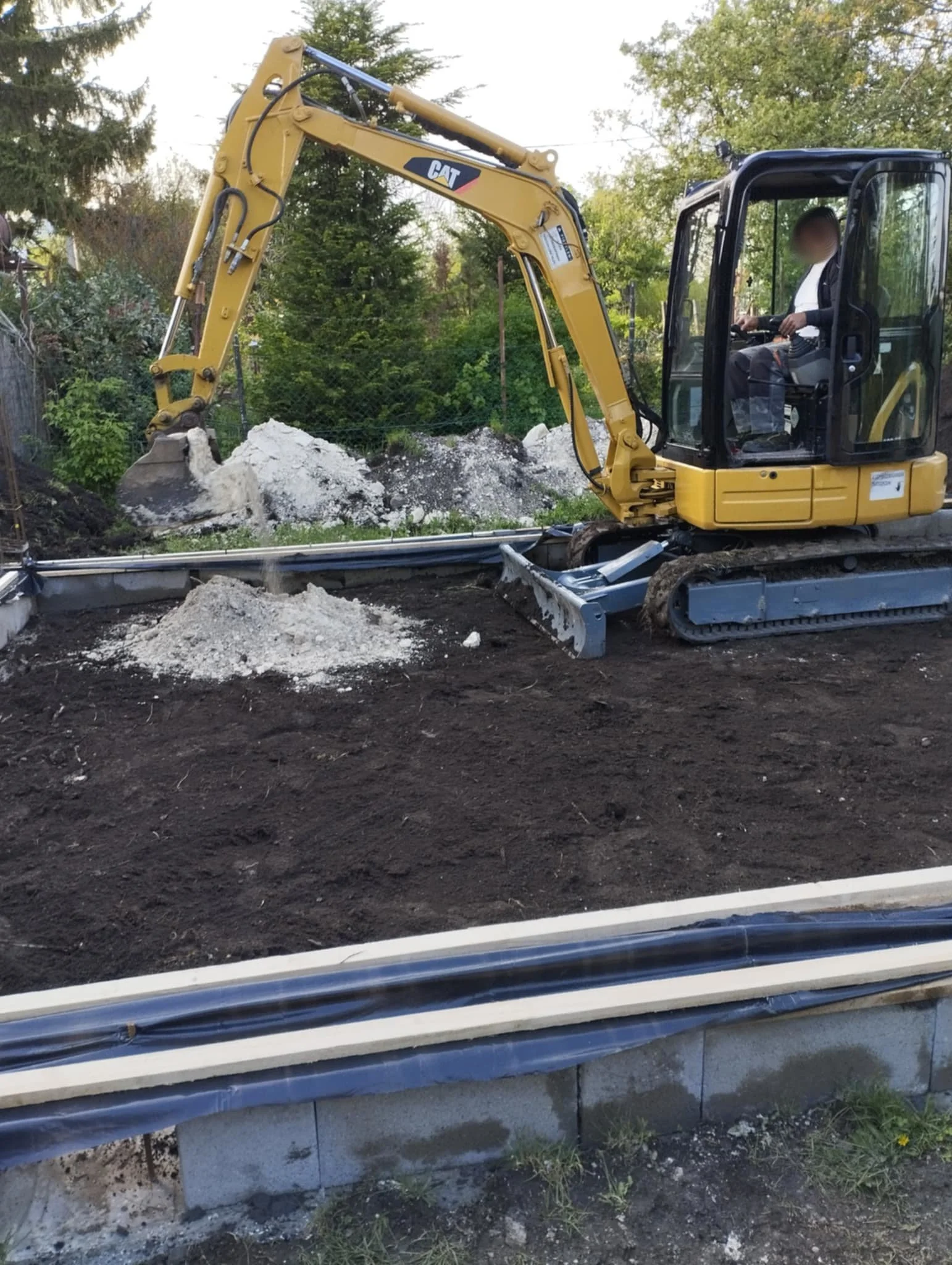 A small yellow Caterpillar excavator operating on a construction site, moving dirt and rocks, with a worker inside the cabin surrounded by green trees.