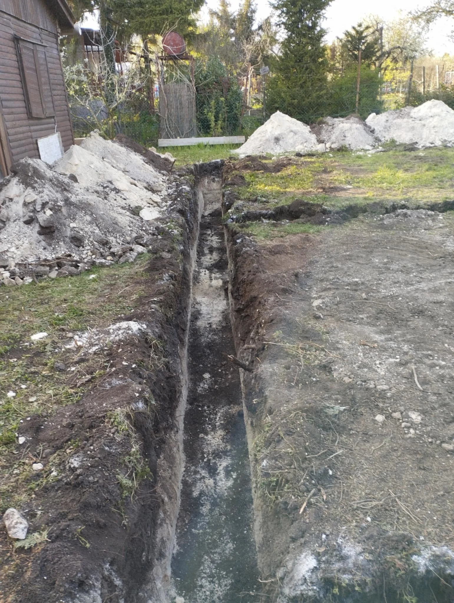 A long, narrow trench dug into the ground outside next to a house, with piles of dirt and gravel nearby, and a garden fence and trees in the background.
