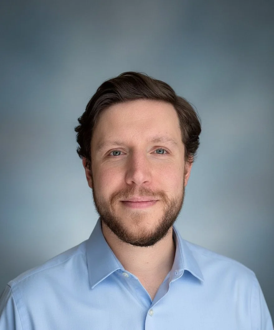 A portrait of a man with brown hair, blue eyes, and a beard, wearing a light blue collared shirt, standing against a neutral gray background.