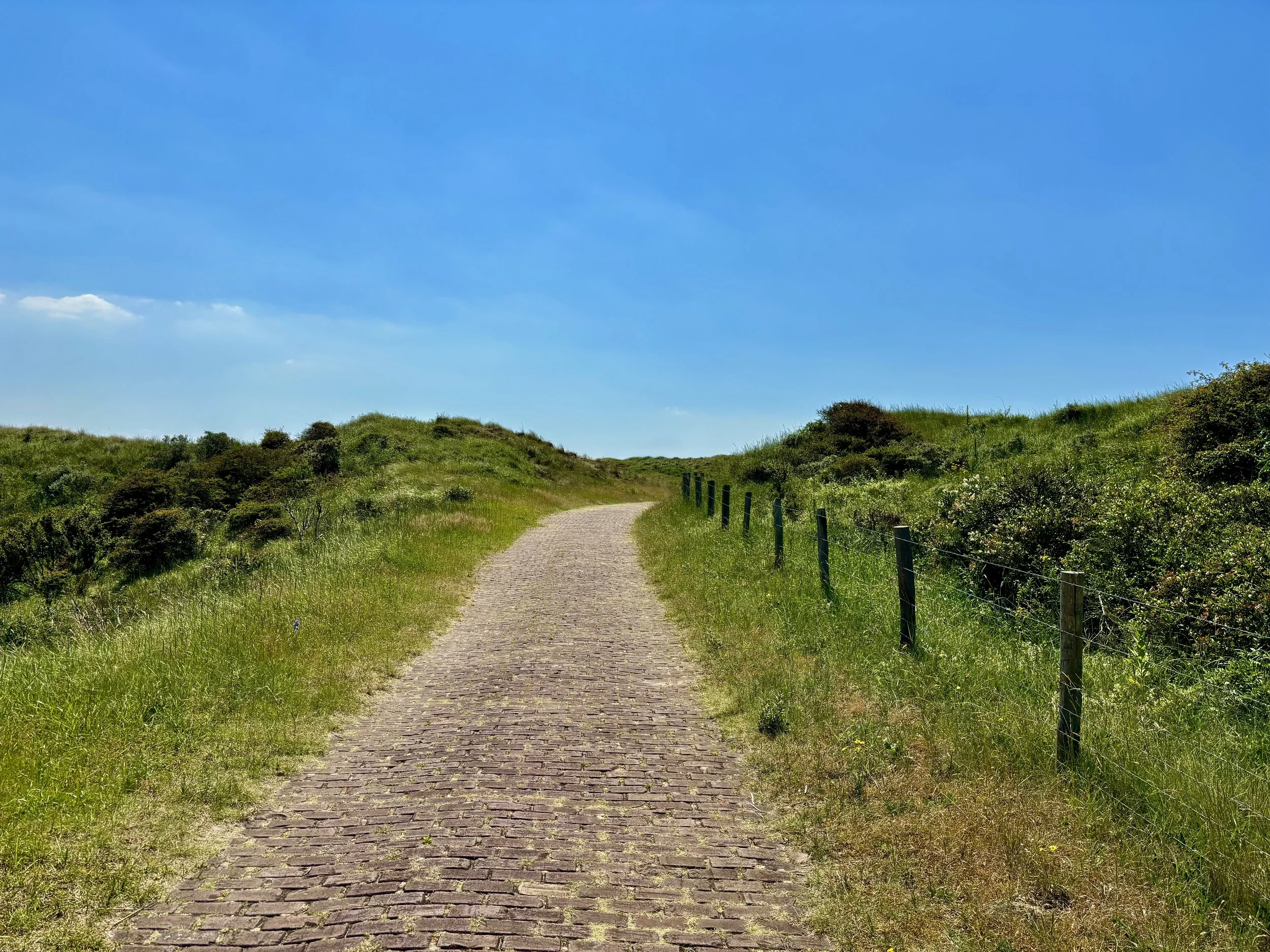 A winding brick path through grassy, hilly terrain under a blue sky with a few clouds.