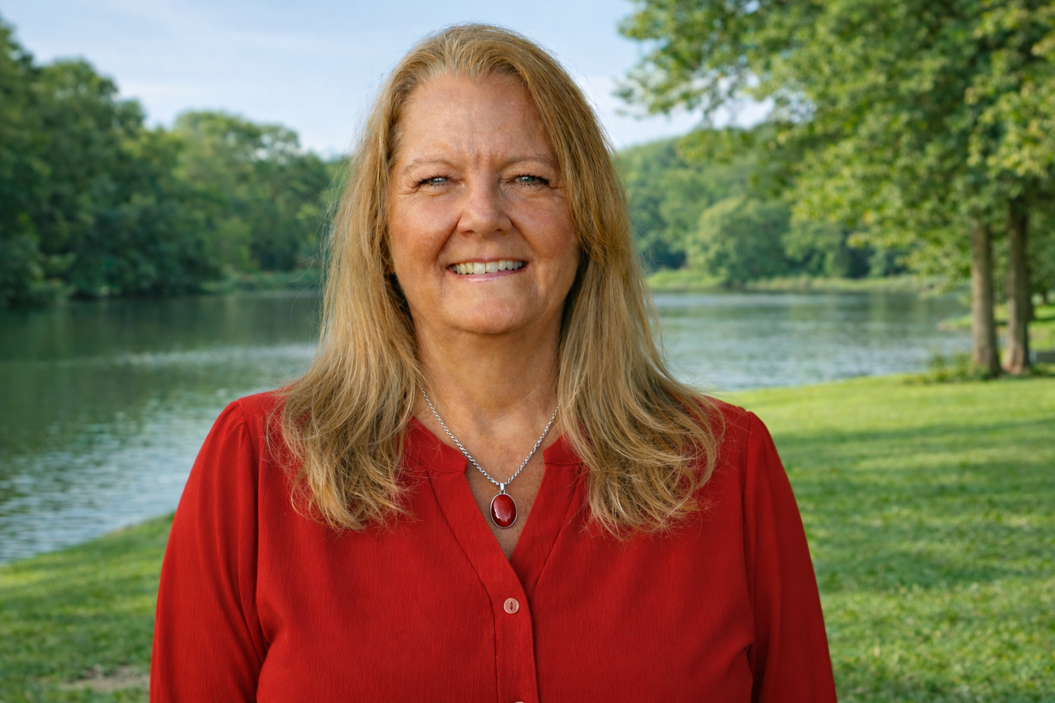 A middle-aged woman with long blonde hair wearing a red top and a silver necklace with a red pendant, smiling outdoors near a river with trees in the background.