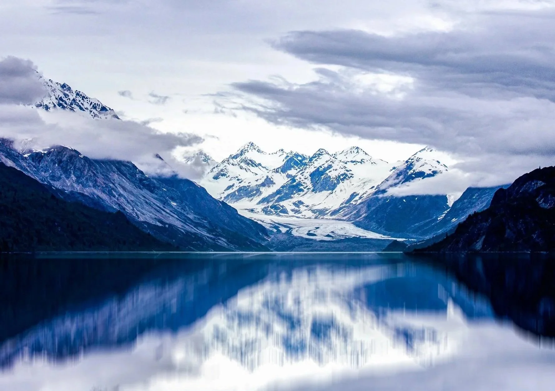 Snow-capped mountains reflected in a calm lake with cloudy sky above. Glacier park, Alaska.